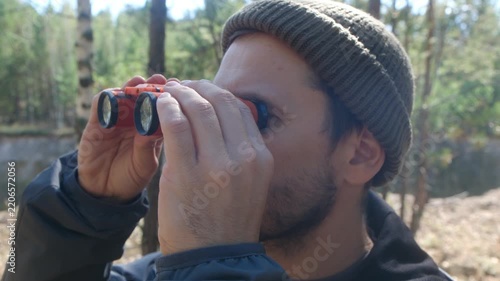 Young male hiker or birdwatcher wearing beanie hat, exploring sunny woods, using pair of red binoculars to observe something interesting in distance. Curious man looking through binoculars in forest