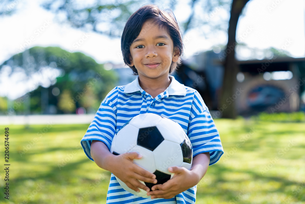 Obraz premium Portrait of happy little Asian boy holding a soccer ball looking at camera in sunny park. Active childhood and outdoor recreation concept.