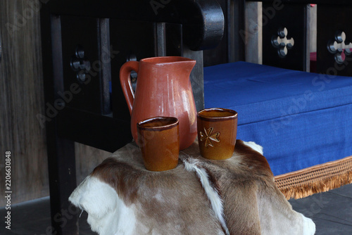 Fotografie Clay pitcher and cups arranged on a goat hide covered stool beside a blue cushioned chair, with a wooden throne visible behind them, recreating rustic medieval hospitality at a fair