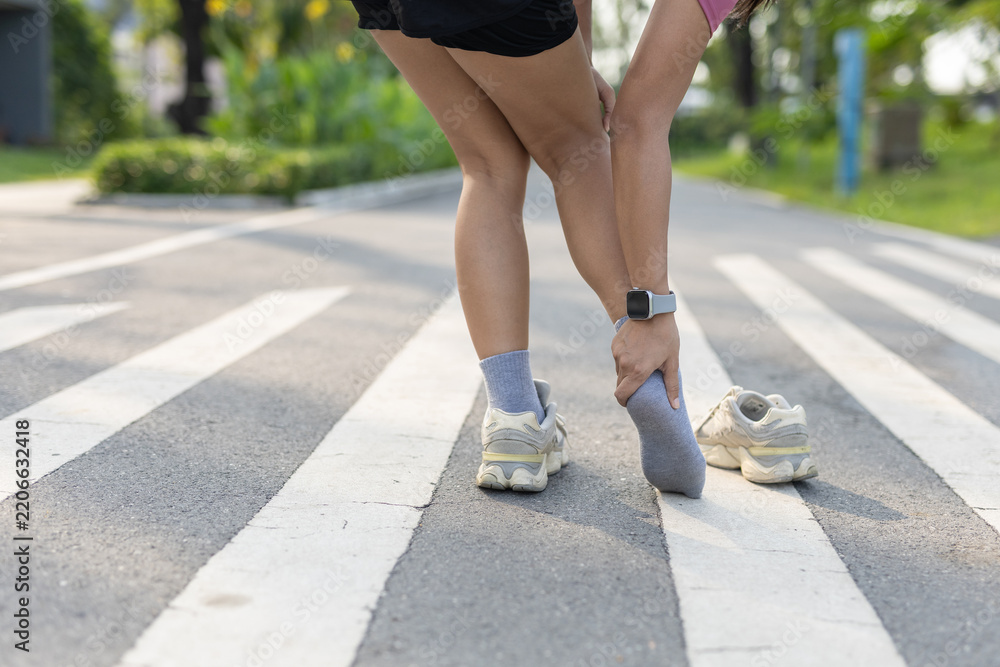 Obraz premium Heel and ankle pain after running women stops on outdoor path, removes sneaker and holding sore foot. Running injury, exercise discomfort, Foot ache.