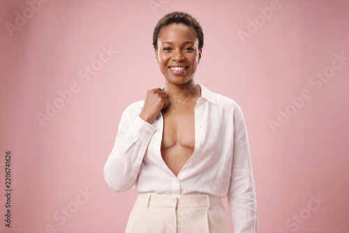 Confident smiling Black woman unbuttoning white shirt against pink background, concept of breast cancer survivor strength and pride.
