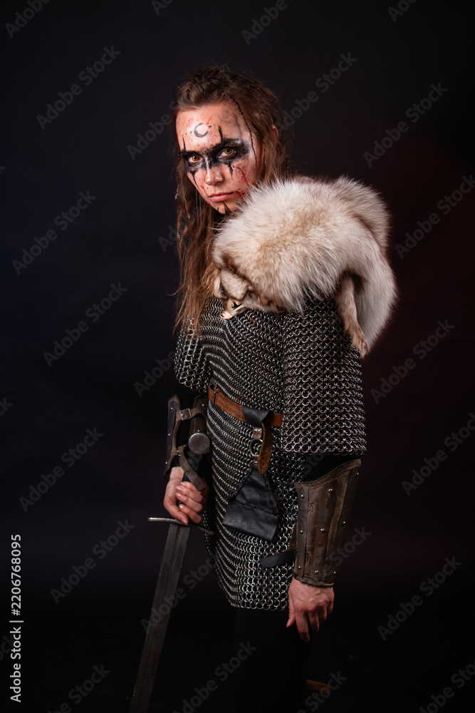 Obraz premium A young Viking woman in fur-trimmed chainmail and war paint, holding a sword, poses against a dark studio background.