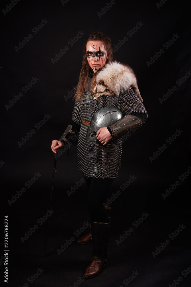 Obraz premium A young Viking woman in fur-trimmed chainmail and war paint, holding a sword, poses against a dark studio background.