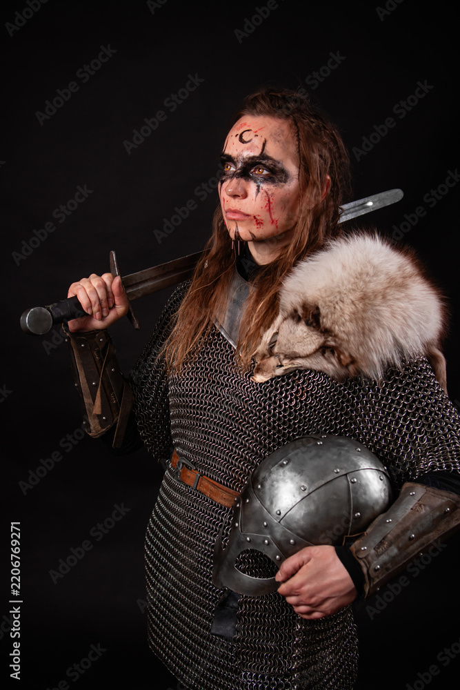 Obraz premium A young Viking woman in fur-trimmed chainmail and war paint, holding a sword, poses against a dark studio background.
