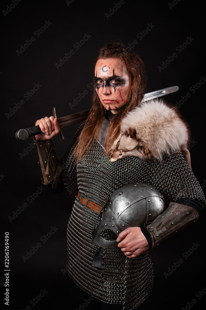 Obraz premium A young Viking woman in fur-trimmed chainmail and war paint, holding a sword, poses against a dark studio background.