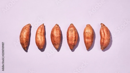 Six organic raw sweet potatoes in a row on a pastel violet background