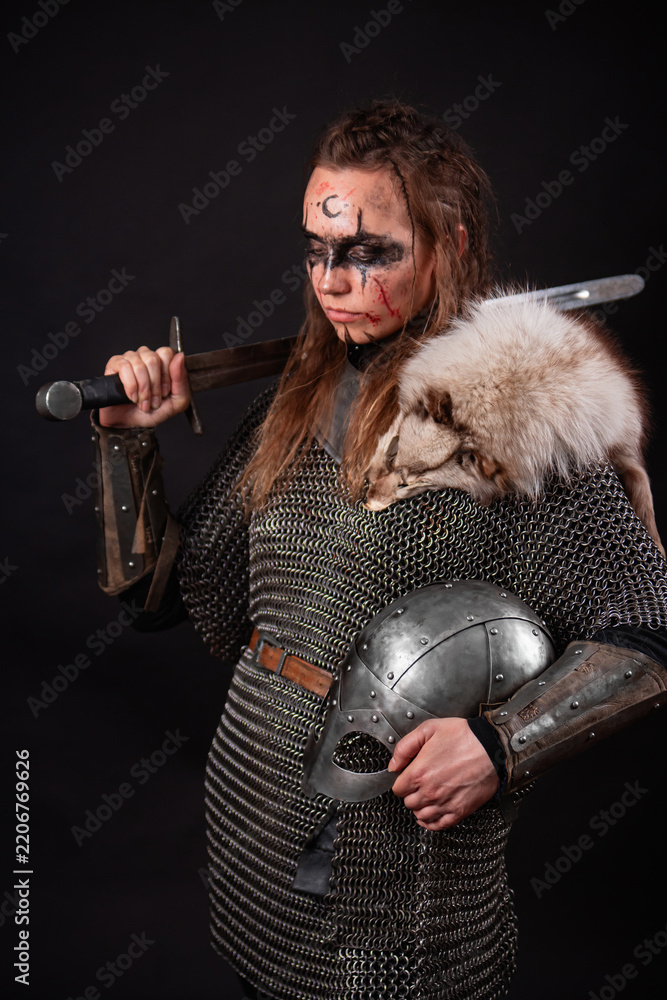 Obraz premium A young Viking woman in fur-trimmed chainmail and war paint, holding a sword, poses against a dark studio background.