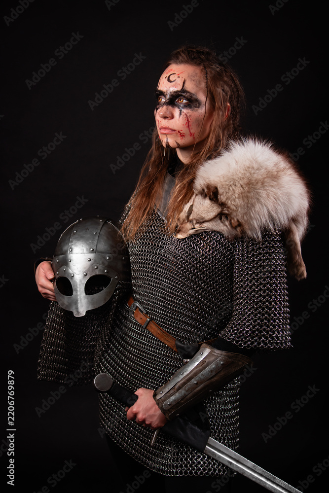 Obraz premium A young Viking woman in fur-trimmed chainmail and war paint, holding a sword, poses against a dark studio background.