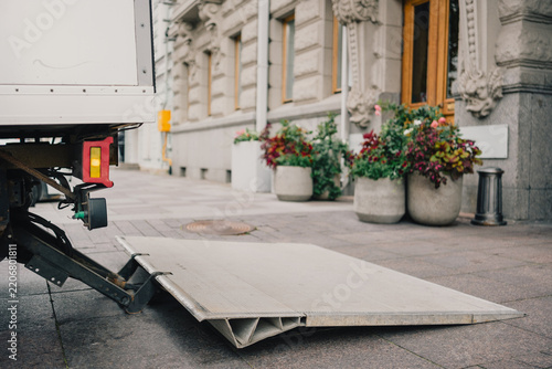 Truck tail lift lowered onto an urban sidewalk beside a building, illustrating commercial delivery, cargo handling, logistics, loading and last mile transportation.