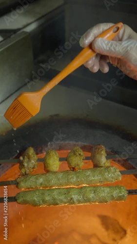 A gloved cook uses an orange silicone brush to baste kebab skewers over a flaming tandoor.