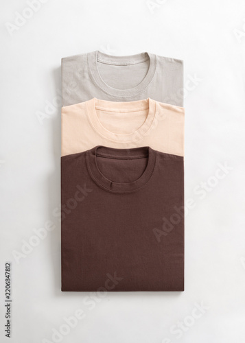 A top down view of a neat stack of three neatly folded cotton t-shirts in earthy neutral shades of grey beige and brown on a clean white background