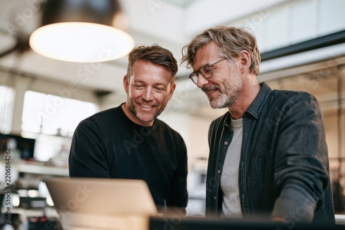 Two male colleagues standing in a bright open office, looking at a laptop screen, smiling and discussing, modern corporate lifestyle