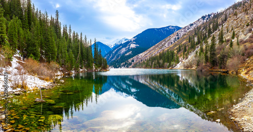 Lake Kolsai in spring. A beautiful spring mountain landscape in Kazakhstan. The blue lake is set against a backdrop of green spruce trees and still-snow-capped mountains.