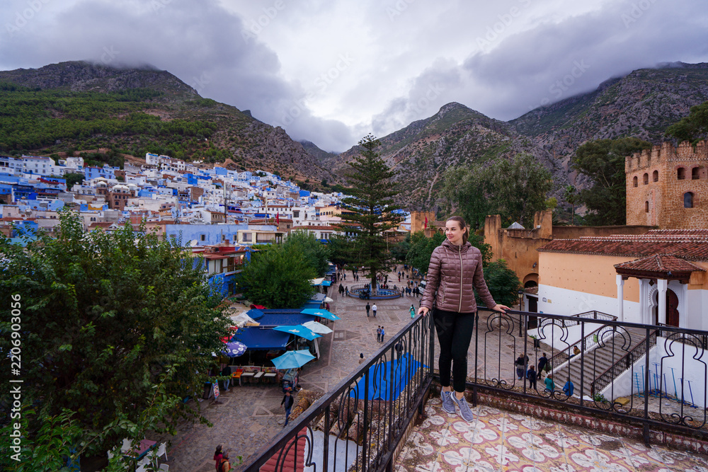 Obraz premium A woman stands on a balcony overlooking a vibrant town with blue-painted buildings, surrounded by mountains. The scene captures a bustling market square below, with people and stalls.