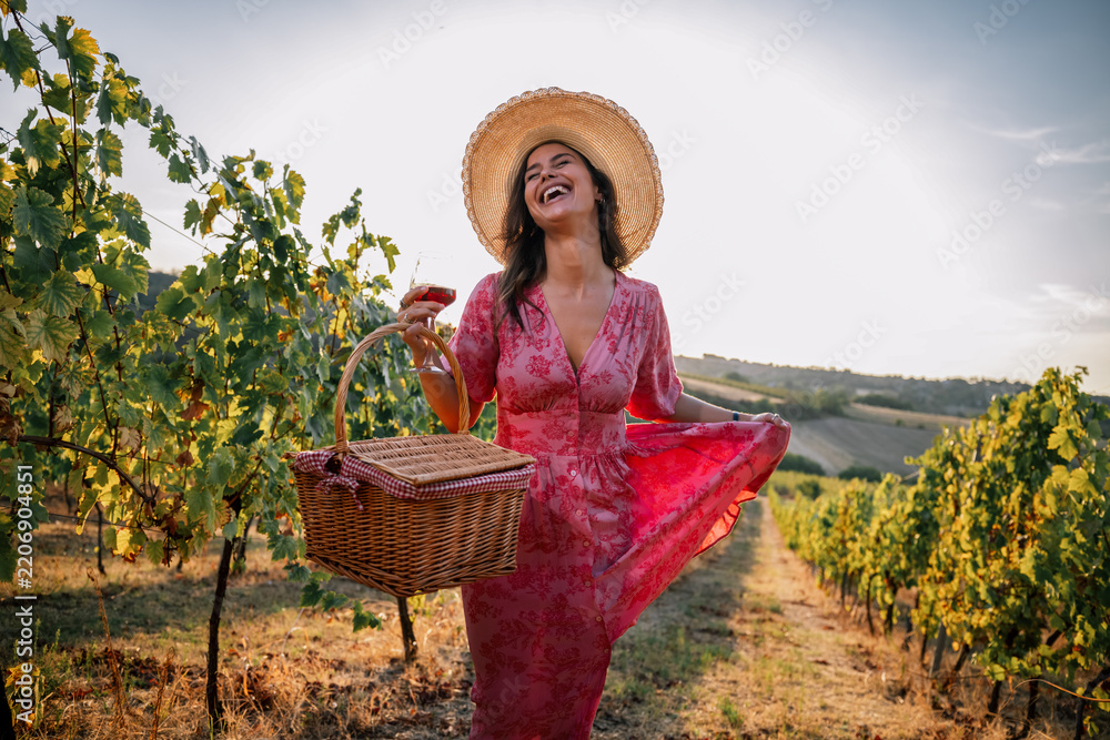 Obraz premium Laughing woman holding picnic basket and wine glass walking in vineyard