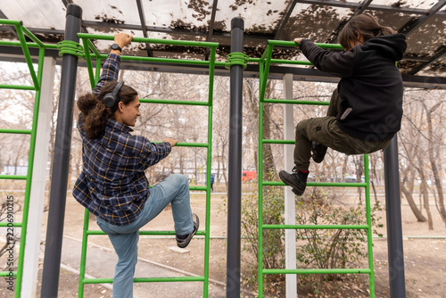 Two young students climbing on green metal bars at an outdoor workout area in a park during autumn.
