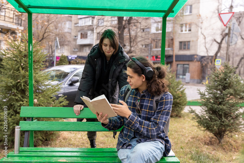 A male student sitting on a bench shows a book to a female classmate standing behind him on campus.