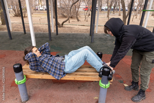 A female student helps her friend with his sit-up exercise on a park bench. Support and student friendship.