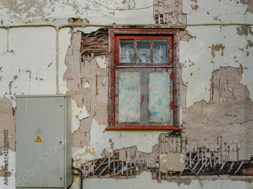Weathered building facade with peeling plaster, an old wooden window, exposed wall structure, and an electrical cabinet.