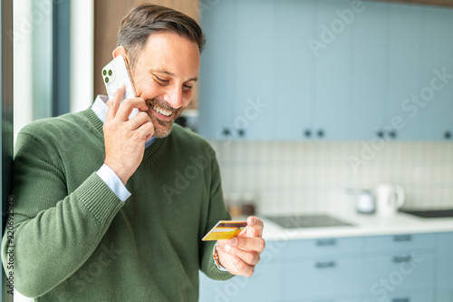 Smiling man making mobile phone call while holding credit card, managing personal finances, making secure online payments, shopping from kitchen