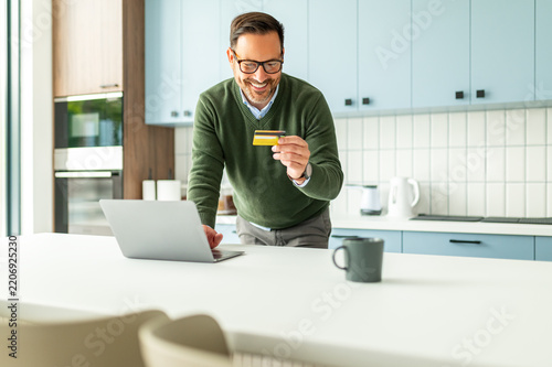 Smiling adult man standing in a modern kitchen setting, holding a credit card and using a laptop for secure online shopping and banking, making an electronic payment