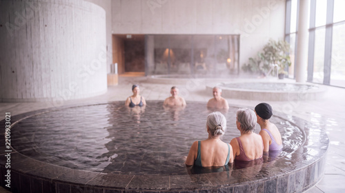 Senior men and women relaxing in steaming indoor thermal pool of modern concrete spa. Mature gray haired bathers in swimsuits enjoy warm water, wellness, health, leisure and holiday calm.