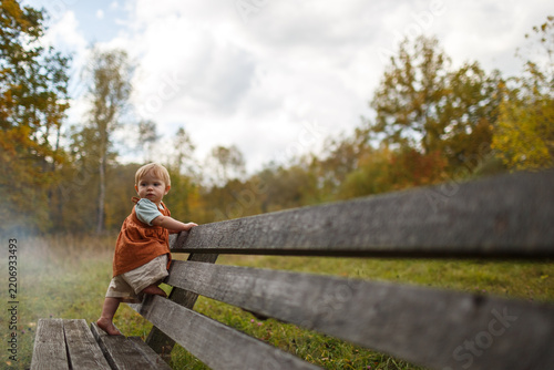Infant baby under one year old standing on a wooden bench in an autumn park outdoors near trees