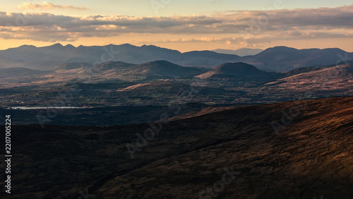 Wide view of silhouetted mountain ridges and rolling hills with a small body of water in the valley at sunset.