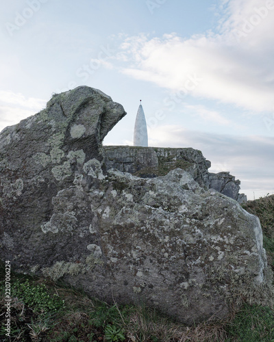 Large lichen-covered rocks stand in the foreground with a white conical monument on a rocky hill under a cloudy sky.