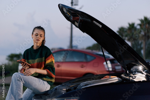 Obraz na plátně A frustrated young woman sits on the open hood of her broken car at dusk, checking her phone