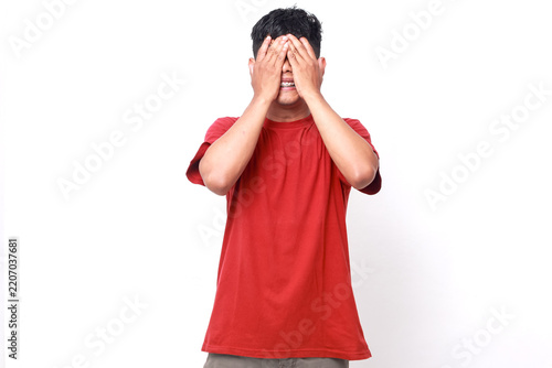 Asian man wearing a red t-shirt covering his eyes with his hands isolated on white background