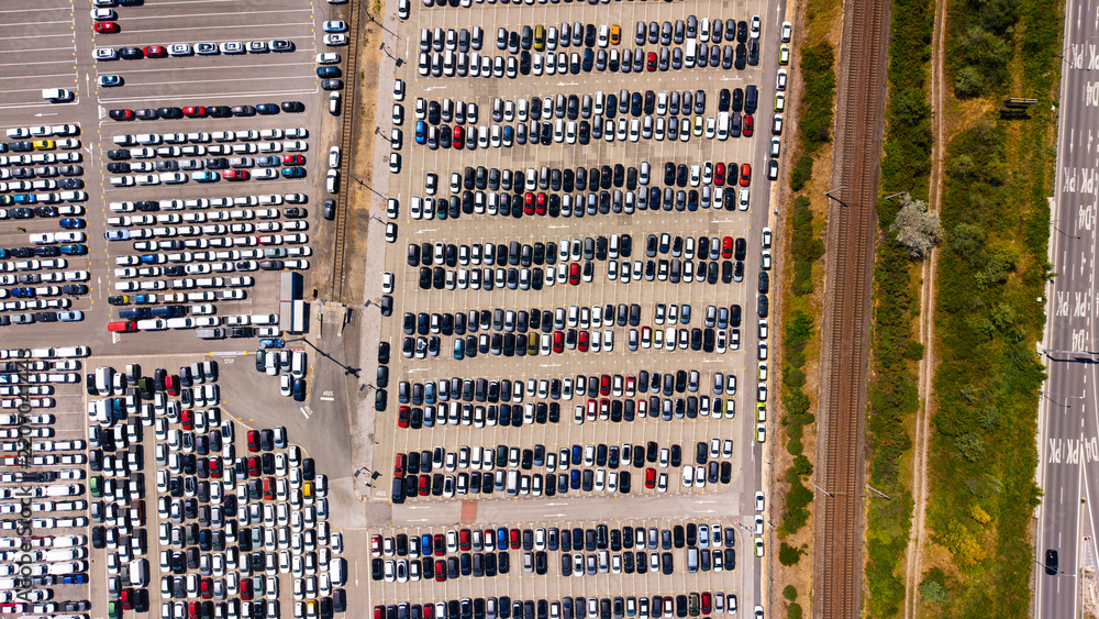 Obraz premium High angle view of organized car rows in an outdoor distribution hub. Hundreds of passenger vehicles sit parked on a paved lot adjacent to railroad tracks.
