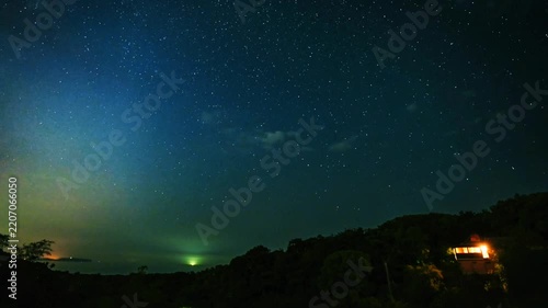 Night sky timelapse revealing millions of twinkling stars, milky way galaxy, moving clouds, and a lit house amidst dark green forest hills, creating a serene, peaceful rural landscape.