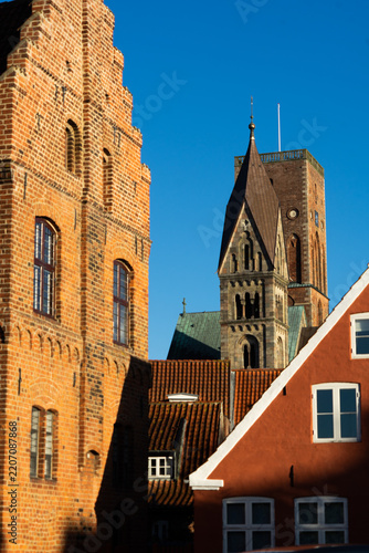 Sunlit red brick step-gabled historic facade in the foreground contrasting with the dark stone tower of Ribe Cathedral in the background under a vibrant blue sky