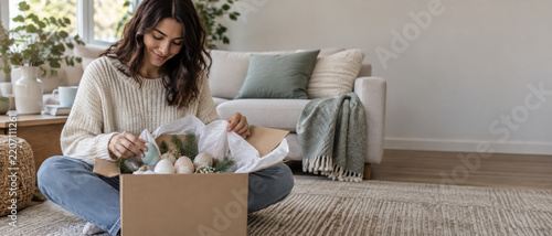 Cozy woman opening holiday gift box with ornaments and evergreen decoration at home, creating warm seasonal celebration scene