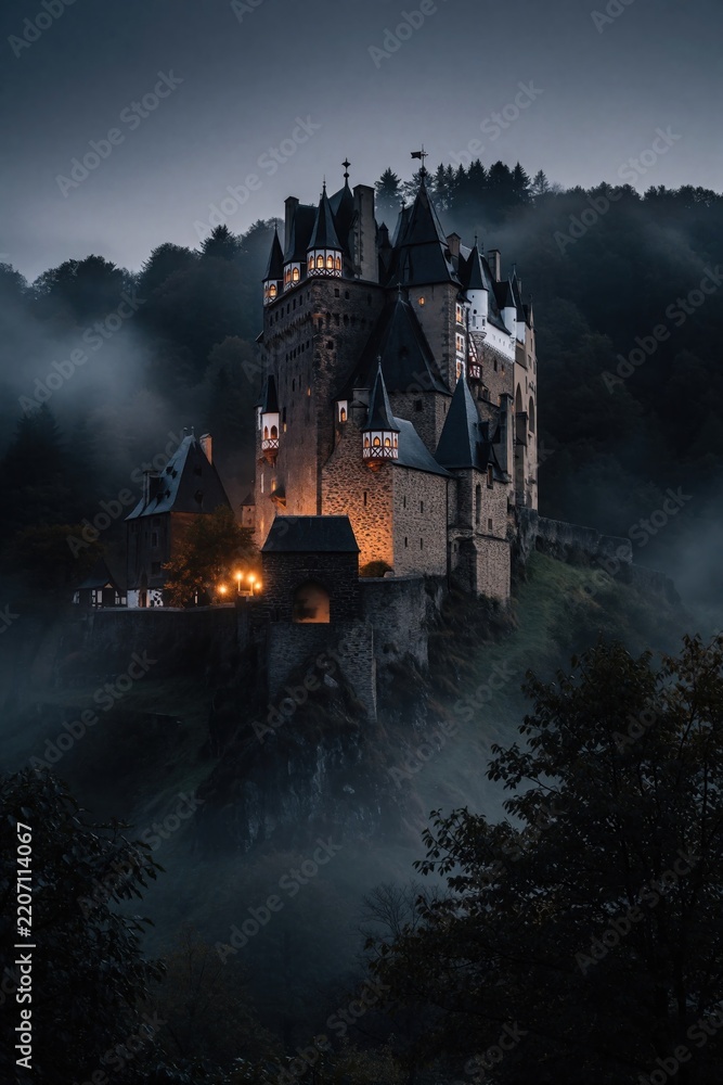 Obraz premium Old medieval castle stands on high rocky hill surrounded by dark foggy forest. Warm lights shine from windows at twilight. Mysterious stone fortress towers over misty woods in moody night atmosphere.