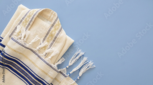 Folded white and blue Jewish prayer shawl tallit with tzitzit fringes on a pastel blue background, flat lay composition with copy space.