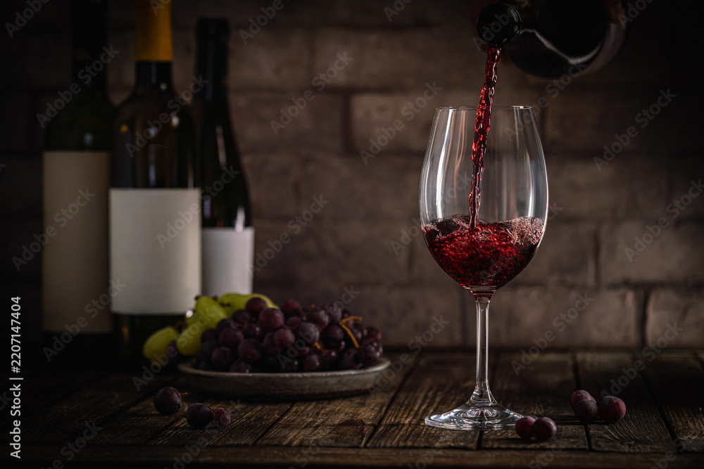 Obraz premium A dark, elegant still life of red wine being poured into a glass with wine bottles and fresh grapes in the background. Perfect for wine-themed compositions
