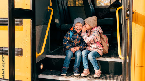 Two young children whispering secrets while sitting on the steps of a yellow school bus together
