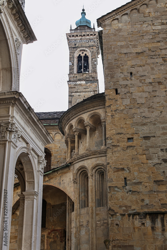 Fototapeta premium Historic stone church tower and ornate arched facade seen from narrow courtyard with weathered masonry, blue cupola, classical architectural details, Bergamo