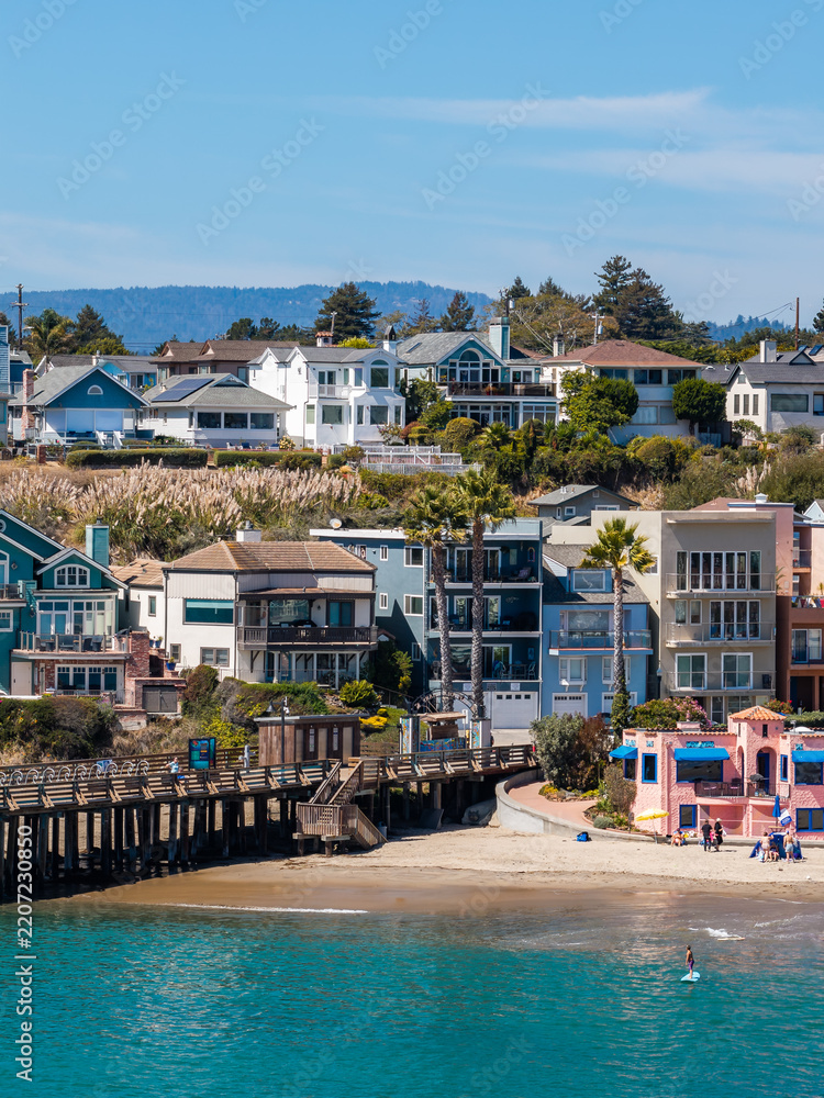 Fototapeta premium Houses and the historic wooden wharf sit beside the sandy shoreline at Capitola Beach in California, where people enjoy water sports and coastal recreation on a sunny day.