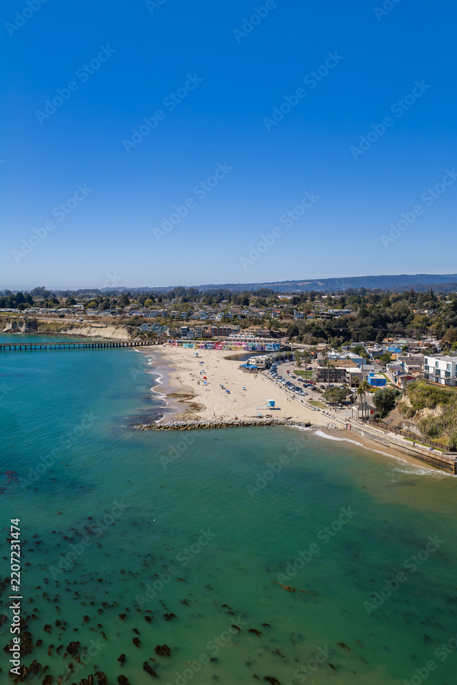 Fototapeta premium Aerial perspective reveals Capitola beach alongside the wooden wharf, beachfront village architecture, shoreline kelp patches, and coastal hills in California under a clear daytime sky.