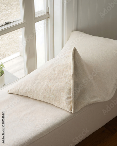 A beige linen throw pillow resting on a long bench seat placed beside a window with white frame in a bright and minimalist room interior space