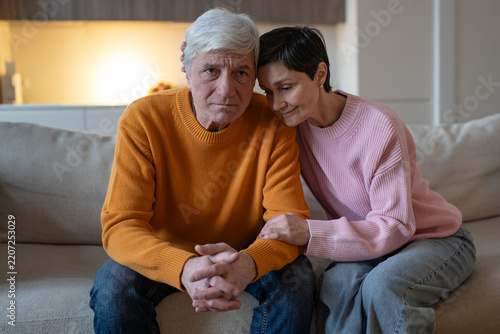 Elderly couple sitting together on couch, compassionate wife embraces worried husband and holds forearm, caring woman gives silent support to distressed partner during difficult life period.