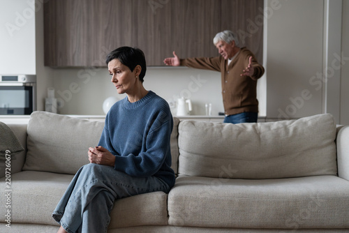Senior marriage crisis and relationship problem, elderly angry husband blaming and arguing with wife, stressed woman sitting on sofa while irritated man shouting behind, emotional distance and divorce