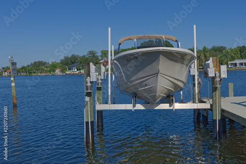 Motorboat stored on a boat lift at a private dock overlooking calm blue water and waterfront homes, conveying leisure, coastal living, summer relaxation and recreational boating on a clear sunny day.
