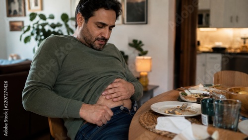 adult man sitting at a dining table after a heavy meal, holding his bloated stomach and unbuttoning his jeans while looking uncomfortable with abdominal pain at home