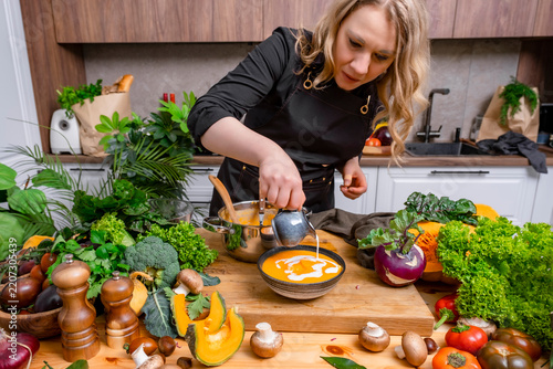 Autumn lifestyle cooking — woman with pumpkin comfort soup. Culinary enjoyment and homemade nutrition concept. Focused female cook in apron pouring fresh cream from jug into bowl with yellow puree.