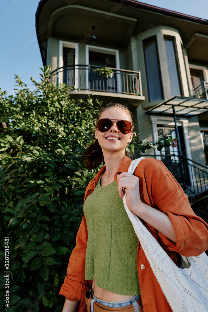 Obraz premium Smiling young woman in sunglasses and orange shirt walking near modern house. Concept of lifestyle, fashion, and summer travel outdoors with happy mood and sunshine.