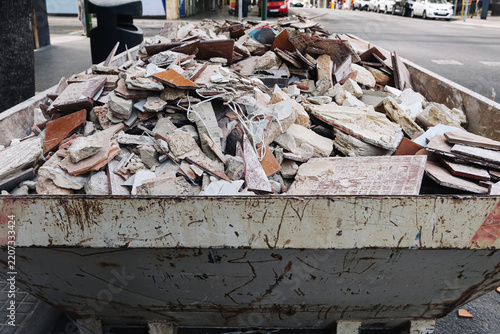 Construction debris in metal container. Broken tiles and concrete rubble. Renovation waste pile. Demolition materials and discarded fragments. Urban cleanup area. Industrial recycling background.